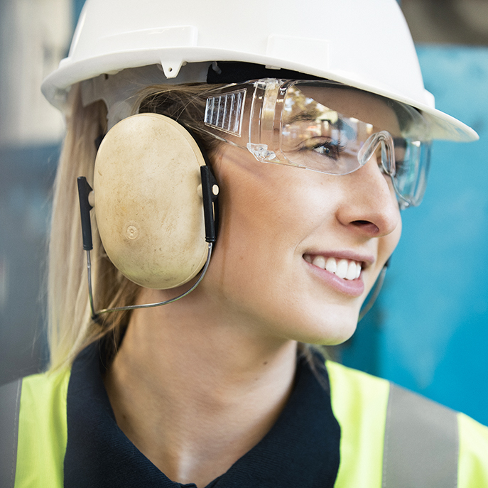 A close-up shot of a young woman engineer at work, she is wearing a hard hat, protective ear muffs, protective eyeglasses​ and a hi-vis jacket.