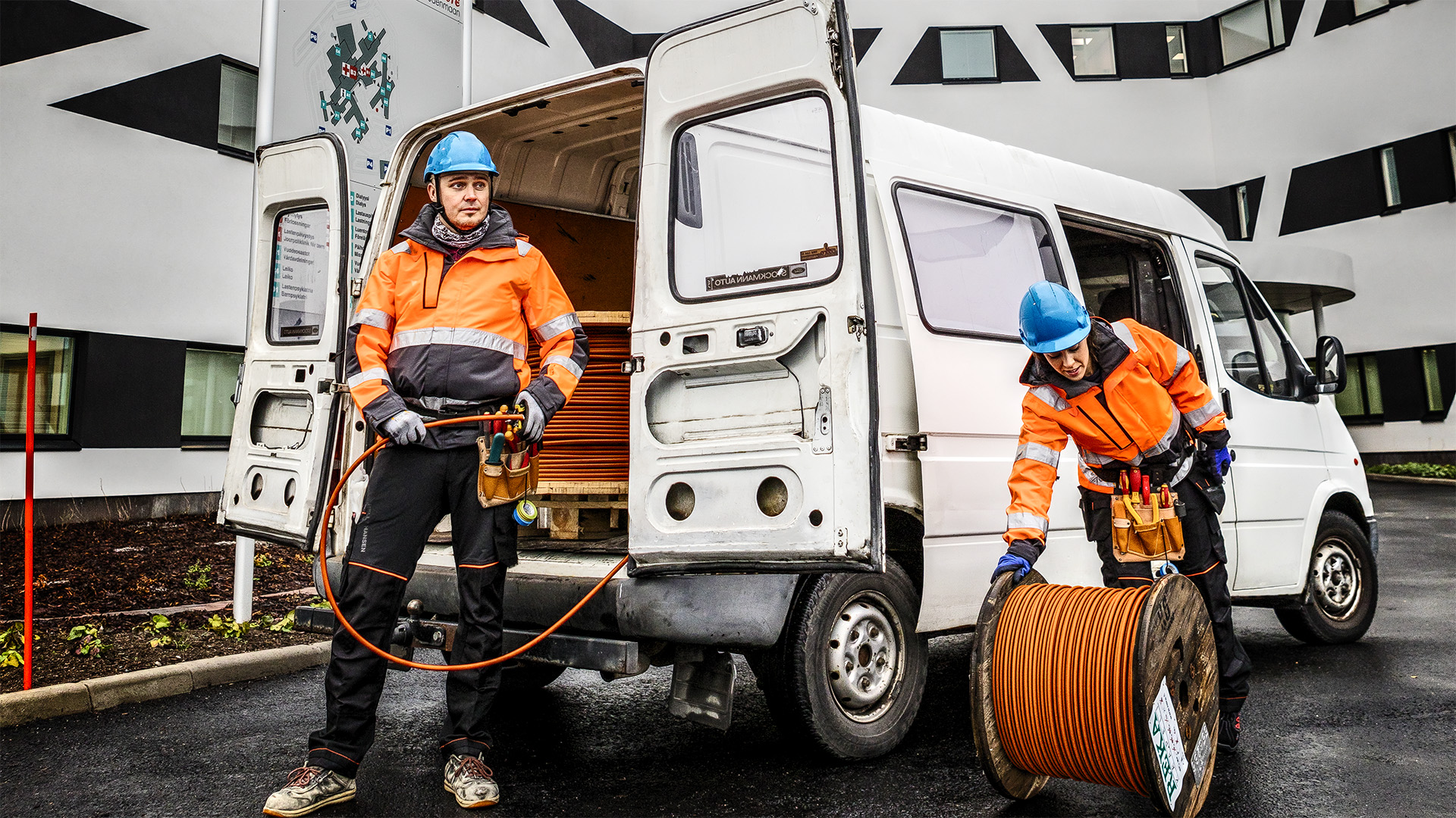 Kuvassa kaksi asentajaa oransseissa työtakeissa ja siniset kypärät päässä ottavat pakettiautosta kelat oranssia FlameRex-kaapelia. Taustalla Hyvinkään valkoinen, moderni sairaala.