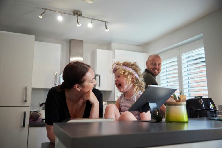 Perhe keittiössä ja mobiililaite keittiön tasolla sekä keittiön sähkölaitteita ja liesihormi sekä spottivalaisin