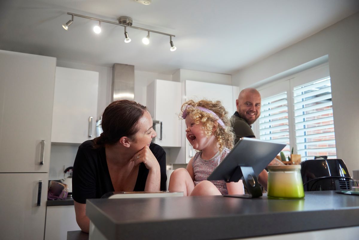 Perhe keittiössä ja mobiililaite keittiön tasolla sekä keittiön sähkölaitteita ja liesihormi sekä spottivalaisin
